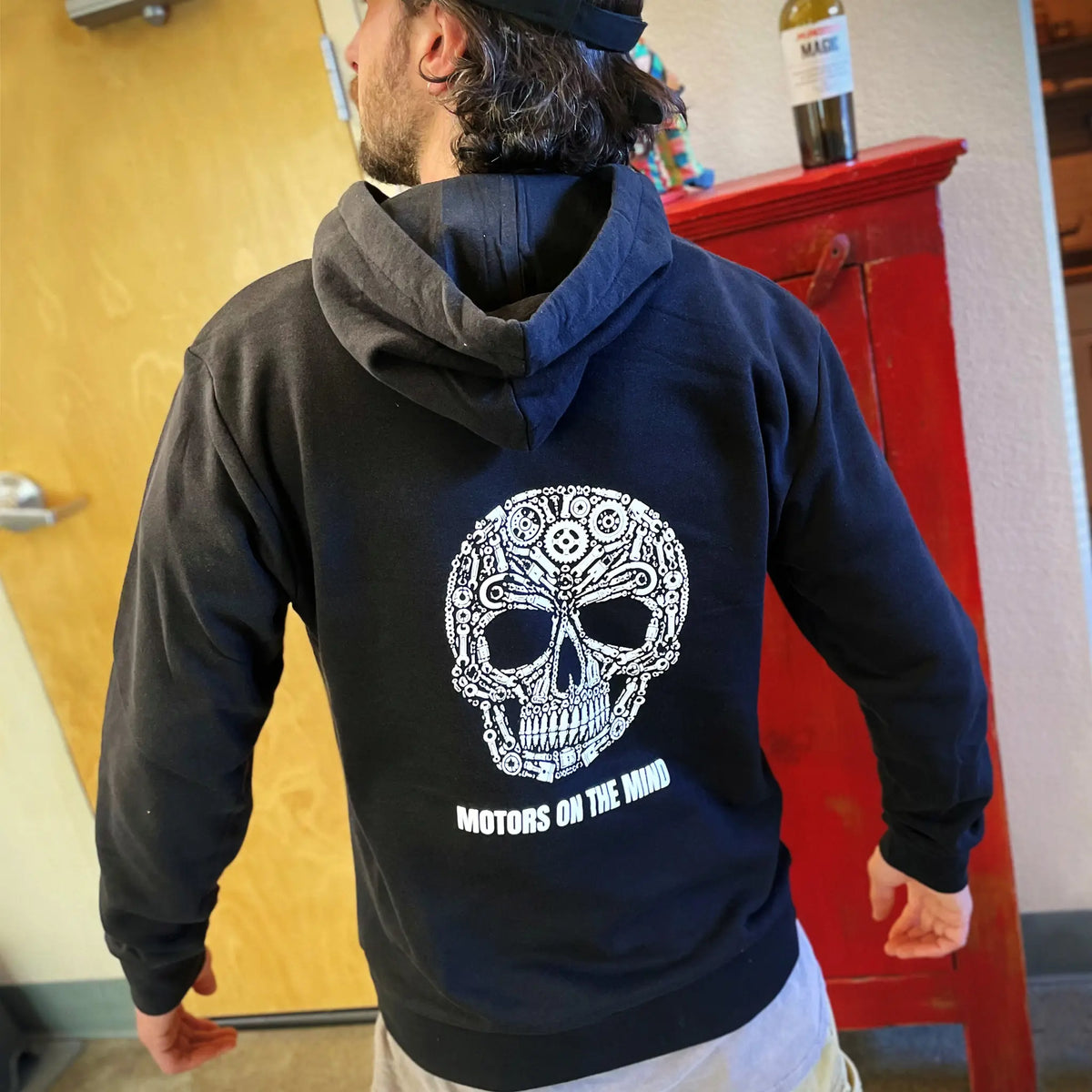 A man wearing a black Motors on the Mind sweatshirt showing the engine-parts skull graphic on the back
