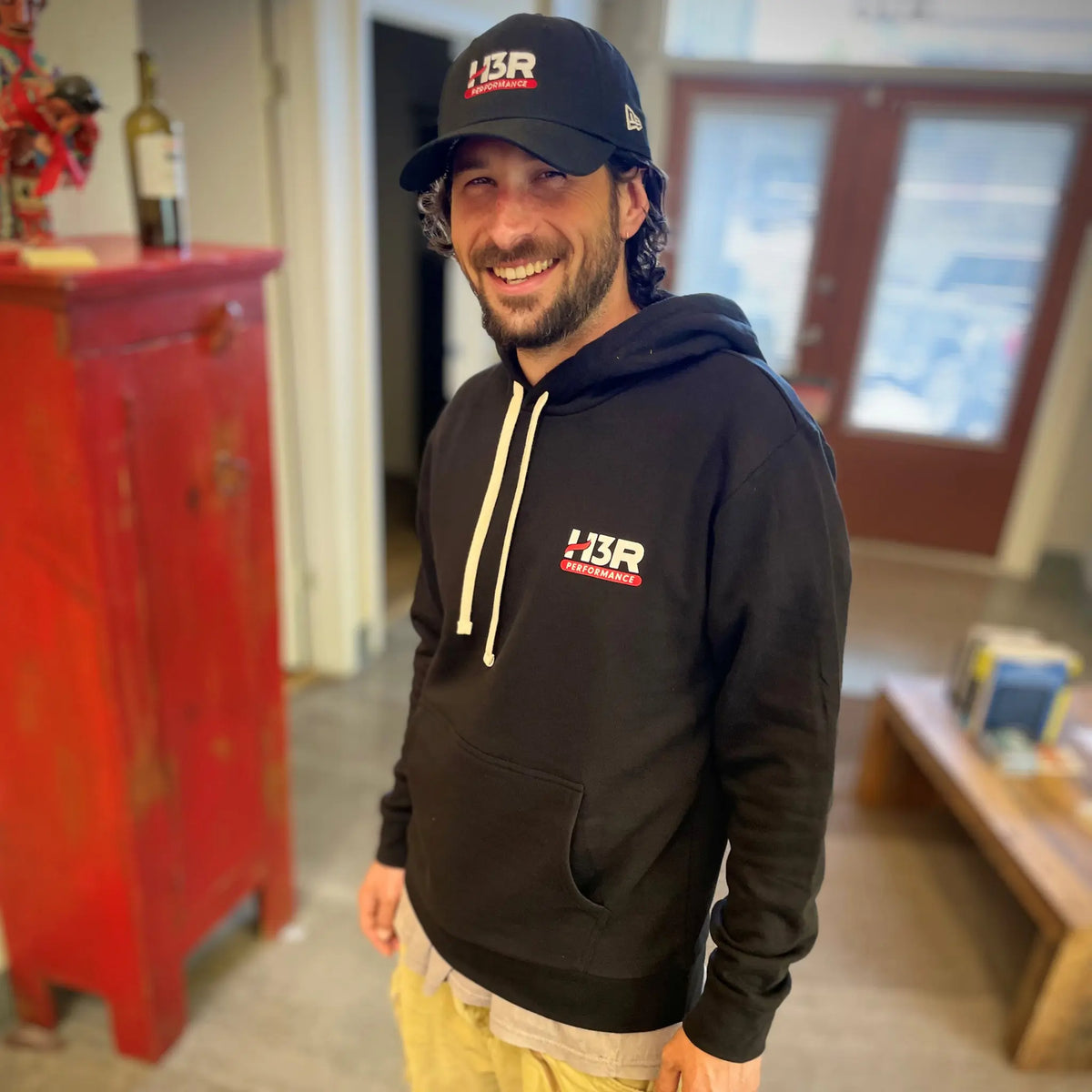 A man wearing a black hoodie with a white and red 'H3R Performance' logo on the chest, a matching black baseball cap, and tan pants, standing in an indoor setting with a red cabinet in the background.