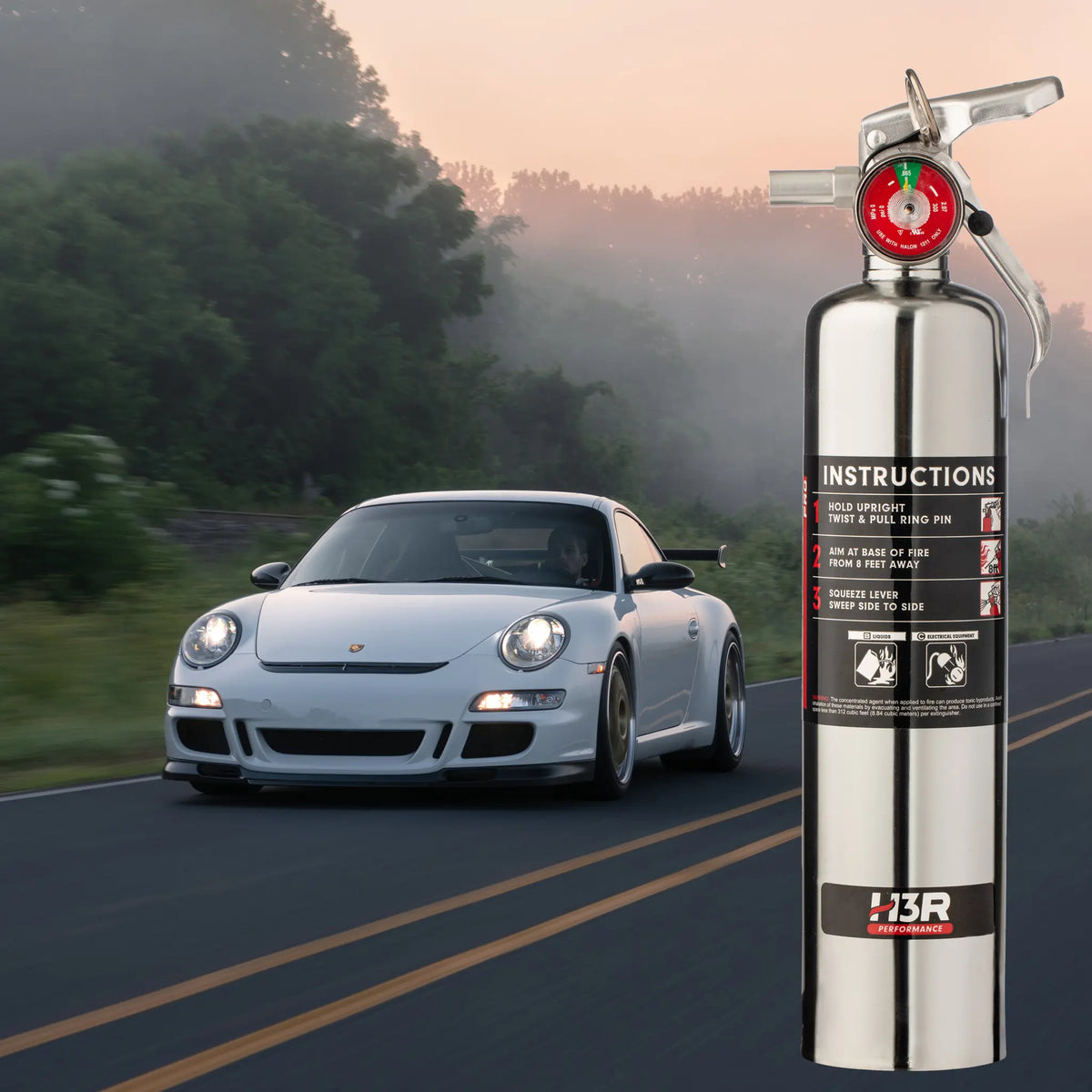 A Chrome H3R fire extinguisher with a red pressure gauge and black instruction label stands prominently in the foreground on a paved road. A white sports car drives past in the blurred background, creating a motion blur effect.