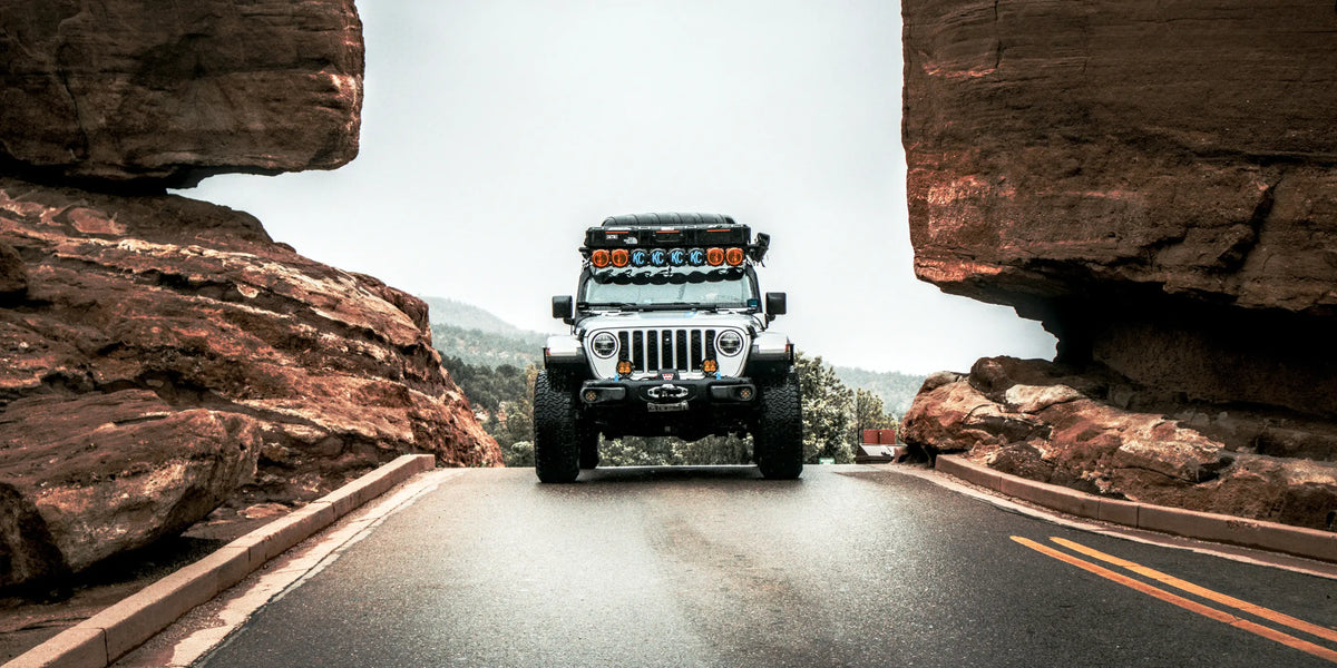A white off-road Jeep with roof-mounted light bar and a tent is driving on a paved road between large red rock formations under an overcast sky.